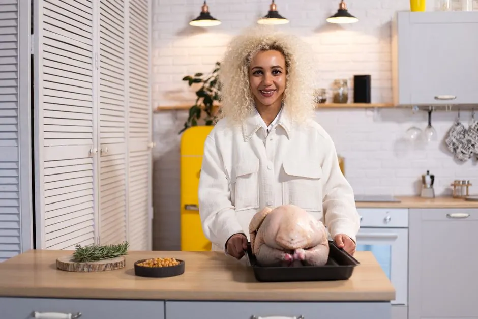 Smiling female looking at camera while standing at counter with uncooked turkey on tray during dinner preparation in modern kitchen